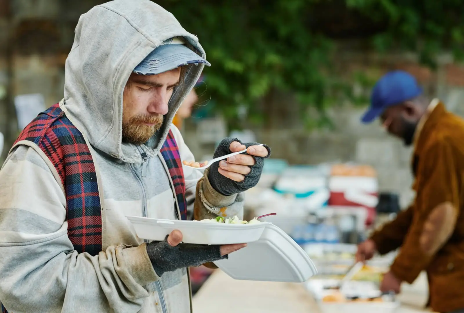 A person in a hoodie eats from a takeout container with a spoon, while another person in the background serves food on an outdoor table.