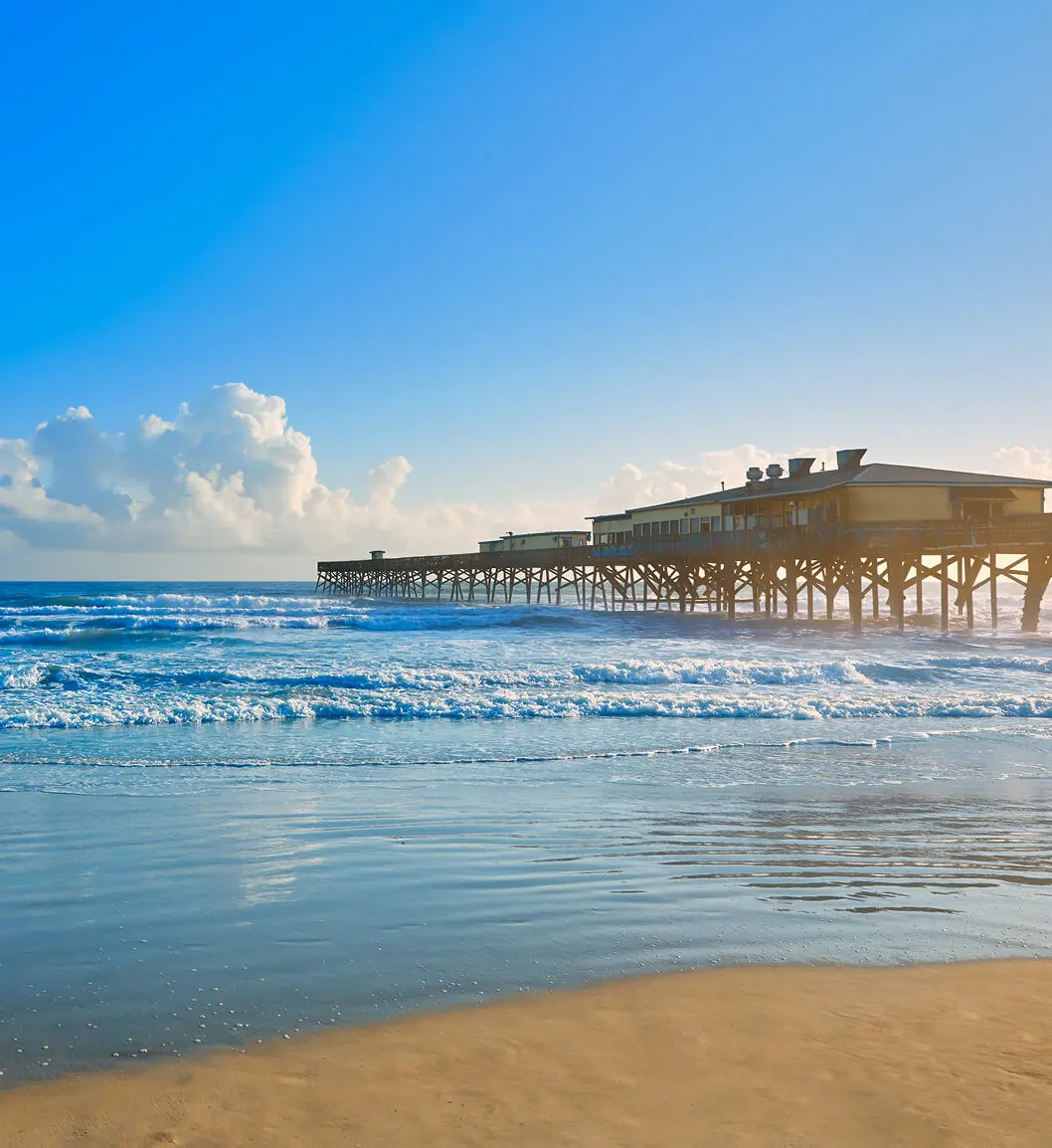 bb22GettyImages-907649378 (1) Wooden pier stretching into the ocean under a clear blue sky, with gentle waves reaching a sandy beach.
