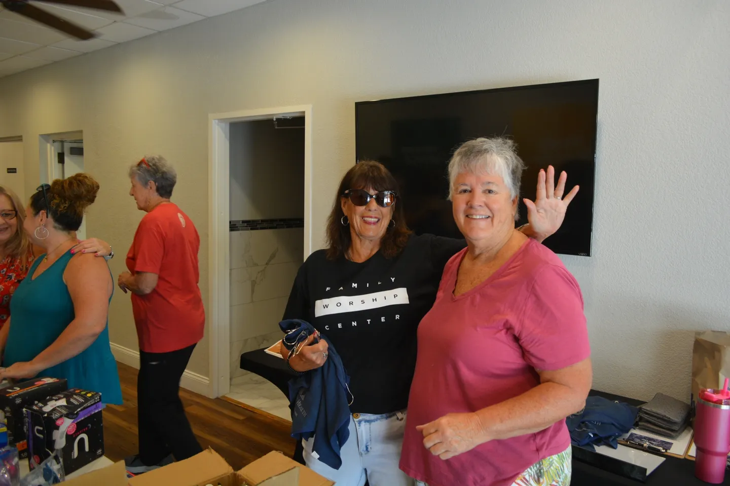 Two women wave to the camera while standing in a room.